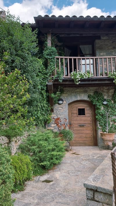 The image shows the exterior of a rustic stone building, likely a house or guesthouse, in Camprodon, Spain. A wooden door with a small, square metal-grated window is centrally located in the stone facade. Above the door is a wooden balcony with a railing, adorned with flowering plants. The building is partially covered in dense green ivy, and the surrounding area features lush vegetation, including bushes and trees. The ground is paved with irregular flagstones, and a stone wall borders the property. The sky above is cloudy with patches of blue. The scene appears tranquil and somewhat secluded, suggesting a rural or mountainous setting within the town.