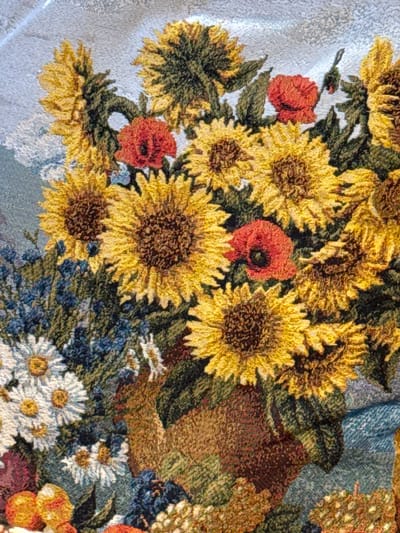 The image shows a close-up of a floral tapestry. The prominent flowers are sunflowers, rendered in shades of yellow and brown. Interspersed among the sunflowers are red poppies and white daisies. Blue and green foliage fills the spaces between the larger blooms. The background is a light blue fabric with a subtle woven pattern. The image is likely a detail shot, focusing on the texture and colors of the tapestry. There are no people, specific actions, or interactions visible in the image. The image does not show the location in the city of Ketoshi, Russia.
