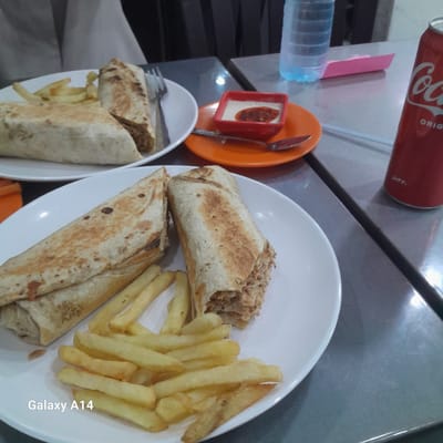 This image captures a casual meal set on a light-colored table, likely indoors at a restaurant or fast-food establishment, consistent with a setting in Nuseirat, Palestinian Territory.

The main subjects are two plates of food, featuring shawarma wraps and french fries. The plate on the right, a white oval dish, holds two large shawarma wraps made with lightly grilled flatbread, revealing a savory filling, likely chicken or meat. Alongside these are a substantial serving of golden french fries. A second, similar white plate is partially visible in the lower-left, also containing a shawarma wrap (one portion appears cut in half) and additional french fries. Forks are placed on both plates.

Accompanying the meal is a small orange dish containing a red rectangular container filled with a white creamy sauce (possibly garlic sauce), topped with a reddish-brown drizzle, possibly chili oil or a spicy condiment. A spoon rests within the orange dish.

Beverages include a prominent red Coca-Cola can, clearly displaying the "Coca-Cola" and "ORIGINAL" logos in white text, with some Arabic script visible on its side. A clear plastic water bottle, partially filled, is also on the table. In the background, a pink item, possibly a napkin holder, is visible. The scene suggests a ready-to-eat meal. No people are visible. The bottom right corner of the image includes a "Galaxy A14" watermark.