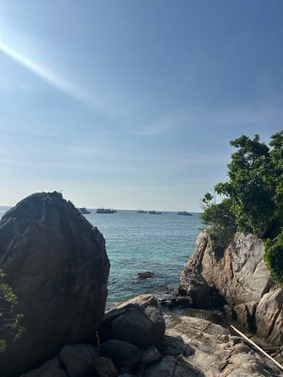 The image depicts a coastal scene in ตำบล เกาะเต่า, Thailand, under a clear, bright blue sky with a visible sunbeam originating from the upper left quadrant. The foreground is dominated by large, dark gray to brown rock formations with visible striations and cracks, partially covered by sparse green foliage, notably on the right side. The midground features calm, clear turquoise-blue ocean water with subtle ripples. Several fishing boats or longtail boats are visible on the horizon line in the distance. No individuals are present in the scene. The overall environment suggests a sunny, natural seascape with rocky shores and open water.
