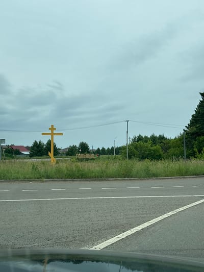 The image shows a roadside view of a large yellow Orthodox cross with three crossbars, indicating it is an Eastern Orthodox Christian cross. To the right of the cross, a sign with Cyrillic lettering, possibly the name of a village or town, is visible. The background features a line of green trees and some houses with red roofs. A paved road with white dashed lines runs in the foreground. The sky is overcast with gray clouds. The location appears to be the outskirts of the village of Забегалово, Russia.