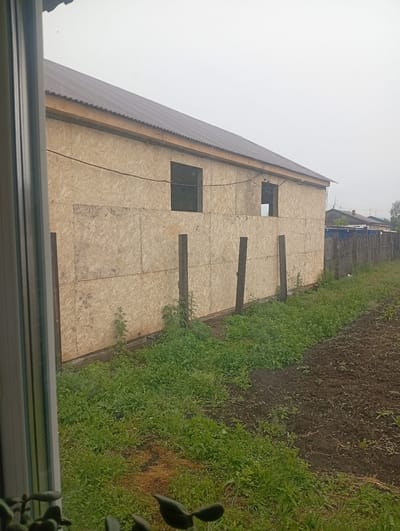 The image captures the exterior of a house under construction in Khomutovo, Russia. The house, clad in oriented strand board (OSB), has a dark, corrugated metal roof. Two rectangular window openings are visible on the side of the building, one boarded up and the other open. Two wooden posts are positioned vertically in front of the OSB cladding. The foreground shows a patch of tilled earth adjacent to an area of dense green vegetation. In the background, a wooden fence runs along the property line, with other residential structures and trees visible beyond it. The sky is overcast and appears hazy. The scene is static, with no people or animals present.