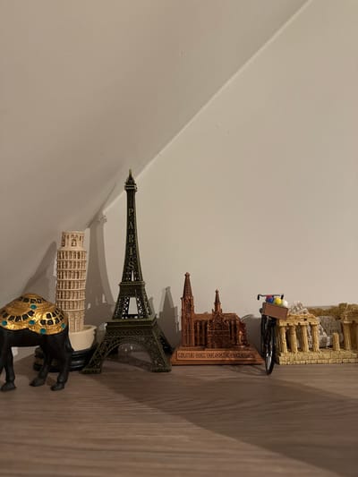 A collection of miniature landmark replicas are displayed on a wooden shelf against a white, slanted wall. From left to right, the objects include a black camel figurine with a jeweled headdress, a leaning tower of Pisa replica, a dark metal replica of the Eiffel Tower with "PARIS" inscribed on it, a copper-colored replica of the Cologne Cathedral labeled "KÖLNER DOM COLOGNE CATHEDRAL", a miniature bicycle with a basket containing colorful balls, and a crumbling replica of ancient Greek columns. A shadow of the Eiffel Tower is cast on the wall behind it. The lighting suggests an indoor environment, possibly a home or shop. The location in the city of Wervik, Belgium, is not discernible from the provided image.