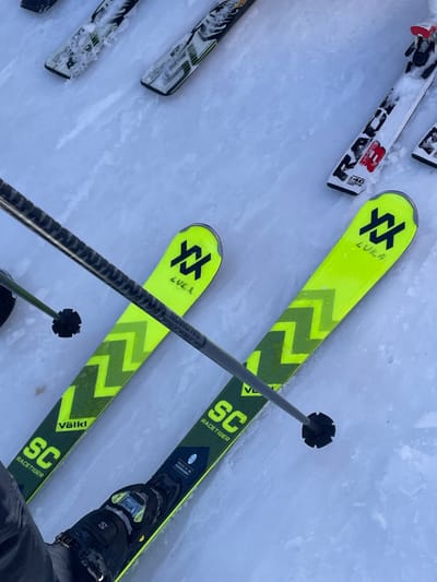 An overhead perspective captures a ski scene in Jahorina, Bosnia and Herzegovina. The primary focus is on a pair of Volkl Racetiger SC skis, predominantly neon yellow with dark green chevron patterns and text, resting on a packed snow and ice surface. The left ski, visible in the lower-left, is connected to a black Salomon ski boot. Both prominent skis display the "Volkl" logo, a distinctive "XX" logo, and the name "LUKA." A gray ski pole, marked "SALOMON," is held diagonally across the image, with its black basket visible near the tip of the left ski. In the background, several other skis are partially visible on the snow, including a white HEAD-branded ski with red "RAB" and "40" markings. The environment consists of uniformly compacted snow or ice, consistent with a ski resort setting.