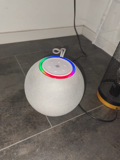 The image shows a white, spherical smart speaker with a colorful light ring around its top controls. The speaker is placed on a dark tiled floor, next to a white baseboard and a corner of a wall. A black power cord is connected to the speaker and runs along the floor. To the right of the speaker, a black mesh waste bin with a wooden base is visible. The scene appears to be indoors, possibly in a domestic setting in Schwäbisch Gmünd, Germany. There are no people present in the image.