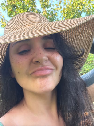 The image is a close-up, self-taken photograph of a young woman wearing a straw hat. She has dark hair and appears to be outdoors, with green foliage and blue sky visible in the background. Her eyes are closed, and her lips are pursed. There are freckles on her cheeks and nose. The image does not contain any information to determine the location within Ароматненское сельское поселение, Ukraine.