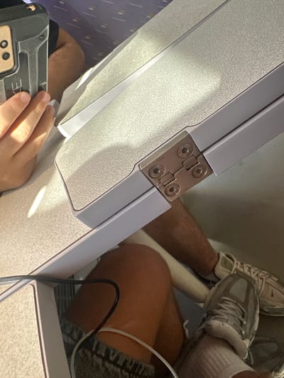 The image depicts a close-up view of a foldable tray table on what appears to be public transportation, possibly an airplane or train, with a person's hand and a phone resting on it. The tray table is light gray with a textured surface and is attached to the seat in front by a metal hinge. Sunlight is streaming onto the table, creating bright highlights and shadows, suggesting it is daytime.
The scene includes parts of two individuals. One person's legs are visible, wearing sneakers and socks, and seated in front of the tray. Their legs are positioned as if resting or extended. Another person's hand is in the foreground, holding a dark-colored electronic device, possibly a phone or a ruggedized case. The device has visible buttons or ports.
The background shows glimpses of seats and the interior of the vehicle. The patterned blue and gray upholstery of the seats is visible, along with the textured surface of the tray table and the metal hinge. The overall impression is one of travel, with a focus on the personal space around the tray table. There is no explicit indication of the time of day other than the sunlight. No weather cues are apparent. The location context of Narbonne, France, does not provide specific visual cues within the image itself.
No legible text is visible in the image, except for what might be part of a logo or branding on the electronic device, but it is too obscured to read. The image captures a moment of stillness during travel, with the emphasis on the tactile surfaces and the presence of personal belongings.