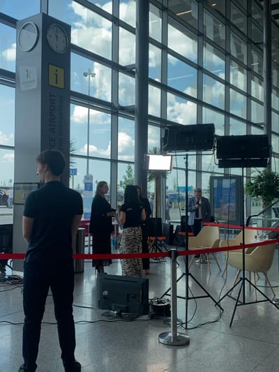 The image depicts a scene inside an airport terminal, specifically in the Katowice Airport in Pyrzowice, Poland. The scene shows a film crew setting up for a shoot, with a cameraman standing in the foreground. Behind the cameraman are three women. Two women are dressed in suits, one in a black suit and one in a white suit. The other woman is wearing a zebra-patterned dress. They are interacting with the camera crew, while a man in a suit is talking on his phone in the background. The scene is lit with several large lights on stands. There are chairs and a table set up for an interview, and a large glass window behind the cameraman provides a view of the exterior of the airport. The setting is clearly an airport terminal, with signage indicating the location as Katowice Airport. The environment suggests the film crew is preparing for a promotional video or media shoot at the airport.
