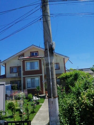 The image depicts a two-story house with a light beige and dark brown facade, situated under a clear blue sky. The house has a red-tiled roof and several windows, some of which are multi-paned. A concrete pathway leads up to the entrance. The front yard is well-maintained with a green lawn, various plants, and flowering bushes. A white van with a Mercedes-Benz logo is parked to the left of the house. A utility pole with numerous wires is prominent in the foreground, partially obscuring the view of the house and its surroundings. The scene appears to be in Tuzla, Romania, as indicated by a number "3" visible on a fence in the bottom right corner, consistent with local street signage.
