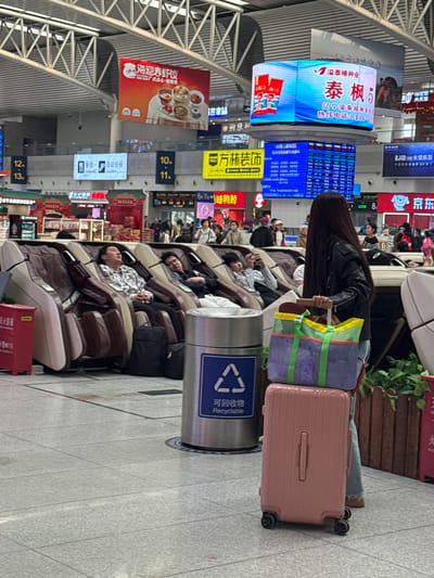 The image shows the inside of a transportation hub in Huanggu District, China. There is a row of reclining massage chairs in the foreground with people relaxing or sleeping in them. The chairs are brown and cream colored. Behind the chairs are a variety of advertisements and signs in Chinese. A woman with long black hair stands near a pink suitcase and a tote bag. There is a recycling bin to her left. Above the massage chairs and to the left is a multi-story display of digital advertisements and notices. The ceiling is a metal framework.
