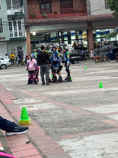 The image depicts a public square or plaza in Timaná, Colombia, during daylight hours. In the midground, a group of approximately five individuals is engaged in what appears to be an inline skating training session. A male adult, wearing a black long-sleeved shirt and grey pants, stands facing away from the camera, seemingly instructing or coaching. Four individuals, primarily children or young adults, are equipped with inline skates, helmets, and protective pads (knee and elbow pads). Two of these individuals on the right wear matching blue, green, and yellow full-body suits, one displaying "TIMANA" on the chest. Another individual on the left wears a pink long-sleeved shirt and black shorts with pink knee pads. A smaller individual is partially obscured behind the instructor. Several bright green conical markers are distributed on the light grey concrete pavement around the group, suggesting a practice area.

In the foreground, to the lower left, a person's foot is visible, wearing a black sneaker with "NEKA" printed on its side. Adjacent to the foot is a neatly stacked pile of approximately twelve identical bright green conical markers. The ground consists of a patterned pavement, featuring light grey concrete squares bordered by reddish-brown brickwork.

The background reveals a bustling urban environment. On the left, a multi-story building with light green walls and white-railed balconies is visible, with a partial sign reading "QUE". To the right, another two-story building features a red-tiled roof overhang, a red brick upper facade, and large ground-floor storefronts displaying a checkered pattern of blue, white, and green panels internally. A street separates these buildings from the plaza. Vehicular traffic includes a grey sedan on the far left, a motorcycle, a black utility vehicle, and a blue car on the right. Additional pedestrians are visible in the background.