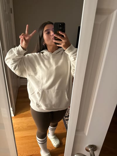 A young individual is captured in a full-body mirror selfie, standing indoors in what appears to be a residential hallway or room within the city of Princeton, United States. The person, with long dark hair, is wearing a light beige oversized hooded sweatshirt with a kangaroo pocket, dark brown leggings, white crew socks with two black stripes, and light green Crocs clogs. They hold a dark-colored smartphone with a prominent camera bump (suggesting an iPhone Pro model) in their right hand, obscuring their face partially. The phone case is black with a visible graphic featuring a tiger's head and the text "TIGER SAVAGE," indicative of an association with Princeton University. In one pose, the person's left hand forms a peace sign while their lips are pursed as if blowing a kiss. The mirror reflects a polished hardwood floor, light-colored walls, a white door frame on the left, and a white door with a silver handle partially visible on the right. A dark patterned rug is faintly visible on the floor to the right.