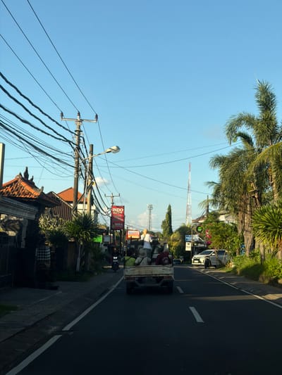 The image depicts a street scene in Kutuh, Indonesia, taken during daylight hours under a clear blue sky. The photograph is oriented vertically. A two-lane asphalt road with a white painted center divider runs from the bottom right toward the center of the frame. A white pickup truck with several passengers in the back is traveling on the road. Overhead power lines are heavily bundled and crisscross above the road. Palm trees and other vegetation are visible along the roadside. Buildings with traditional Balinese architecture, including tiled roofs, are located adjacent to the road. In the background, a tall, slender telecommunications tower is visible, with another slightly more distant, in the center of the frame. Various signs and advertisements, including one for "Indo BISA," are posted along the roadside. A streetlight on a wooden pole is visible on the left side of the road.