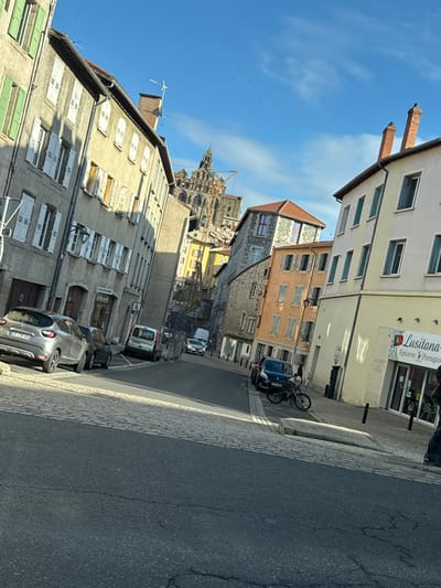 The image depicts a sloped urban street scene in Le Puy-en-Velay, France, under a clear blue sky with sparse white clouds. The perspective is looking uphill, with the prominent Cathédrale Notre-Dame du Puy visible in the distant background, partially obscured by foreground buildings and featuring extensive scaffolding and a large construction crane, indicating ongoing work.
On the left side of the street, a row of multi-story buildings with light grey and beige facades features numerous windows, predominantly with closed white shutters and some green shutters on the leftmost building. One building midway up the left side displays a "COIFFURE" sign. A silver-grey Renault Captur SUV (license plate reads "DF-871-FA") is parked curb-side in the immediate foreground, followed by a dark sedan.
On the right side of the street, another row of multi-story buildings presents varied facades in pale yellow, light orange, and grey stone. The building in the immediate right foreground is yellow and houses a storefront identified by a sign as "Lusitana - Épicerie Portugaise" (Portuguese grocery store). A black mountain bicycle is parked upright on the sidewalk in front of parked dark-colored sedans further up the street. A single individual, clad in dark clothing, is partially visible near the entrance of the "Lusitana" store on the far right.
The street surface in the foreground consists of asphalt, transitioning to cobblestones along the edges and further up the incline. Shadows are cast by the buildings and vehicles, indicating direct sunlight from the left, suggesting morning or late afternoon. Multiple streetlights and poles are distributed along the street. A white utility van and other vehicles are visible driving or parked further up the ascending street.