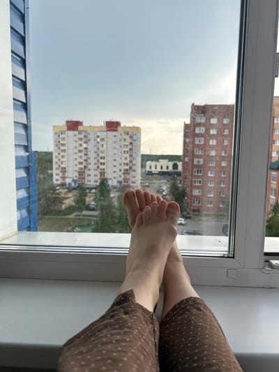 The image shows a first-person perspective of a person's bare feet resting on a white windowsill, with a view of an urban landscape through a window. The feet are crossed, with the toes of one foot resting on the sole of the other. The person is wearing brown pants with a small white polka dot pattern.
In the background, there are several multi-story apartment buildings. The closest one to the left is a light-colored building with red and yellow accents, and multiple balconies. To the right of it is a reddish-brown brick building with many windows. Further in the distance, there are more buildings and trees, indicating a residential area. The sky is overcast and appears to be late afternoon or evening.
The window frame is white, and there is some dust and possibly streaks on the glass. The windowsill itself is also white and appears to be made of plastic. The overall impression is of a quiet, relaxed moment at home, looking out at the city.
The specific location in the city of Солнечный, Russia is not explicitly indicated in the image itself, but the architectural style of the buildings and the presence of trees suggest a setting typical of Russian residential areas.
