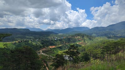 The image depicts a panoramic view of a lush, mountainous landscape in the vicinity of Tu Póh, Vietnam. Rolling hills covered in dense green vegetation and scattered pine trees dominate the scene. Several small clusters of buildings, likely a village, are visible nestled amongst the hills, with red-tiled roofs and white walls. Winding dirt roads cut through the landscape, indicating human presence and access. The terrain is characterized by a mix of steep slopes and flatter areas, with visible terracing in some sections, suggesting agricultural activity. In the background, a series of larger, mist-shrouded mountains recede into the distance under a partly cloudy blue sky. The foreground is filled with tall grasses and more pine trees, creating a sense of depth and framing the expansive view. The overall impression is one of a tranquil, rural environment with evidence of human settlement integrated into the natural beauty of the highlands.