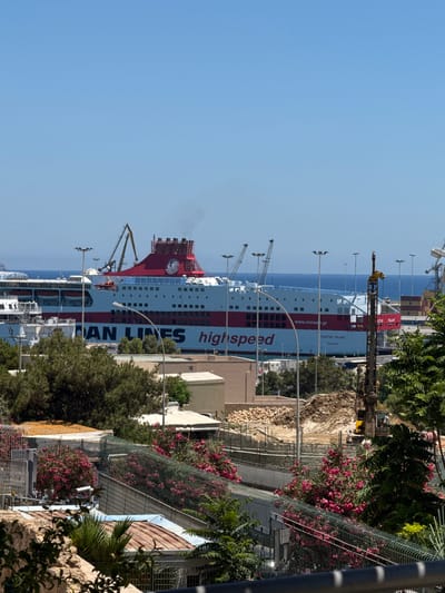 In Heraklion, Greece, a large ferry, identified as "HIGH SPEED" and bearing the Minoan Lines logo, is docked at a port under a clear blue sky. The ferry is predominantly white with a striking red stripe running along its hull and a red superstructure. Smoke emanates from its stack. In the background, industrial cranes are visible, suggesting ongoing port operations. The foreground features lush green trees and flowering bushes, with some metal fencing. A construction site with a pile of dirt and a drilling rig is also present, indicating development or maintenance work. The water is calm and blue, meeting the horizon under the bright sun.