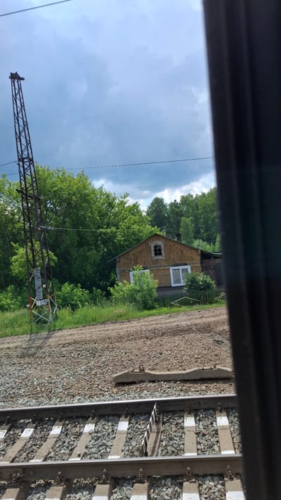 The image depicts a rural landscape with a wooden house and railway tracks. A tall, metal tower with a latticed structure stands to the left of the house, near the railway line. The house is a two-story wooden structure with a visible chimney and a distinctive arched window on the upper floor. It has white-framed windows on the lower floor. A small green fence surrounds the front of the house. The area is overgrown with trees and vegetation, indicating a natural or semi-natural environment. The foreground shows railway tracks with ballast and concrete sleepers. The sky is cloudy, suggesting overcast weather. The view appears to be taken from a moving train, as indicated by the slightly blurred dark vertical element on the right side of the frame, possibly a window frame or part of the train's interior. The location is identified as Сулемка, Russia.