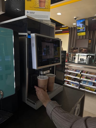The image depicts an interior scene, likely at a gas station convenience store, as suggested by the stocked shelves of snacks and beverages visible in the background. In the foreground, a person's hand is shown reaching for or holding a paper cup under a beverage dispensing machine. Two paper cups are visible on the machine's drip tray, with one appearing to be actively filling with a beverage. The machine itself is metallic and modern, featuring a touchscreen interface. On the wall behind the machine, there are advertisements and signage. One sign, in Russian, appears to promote a "Family Team" initiative and a coffee brand, "ZERNO." The overall environment is clean and well-lit, suggesting a commercial setting. The specific location in Kochubeyevskoye, Russia is inferred from contextual information not directly present in the image itself.