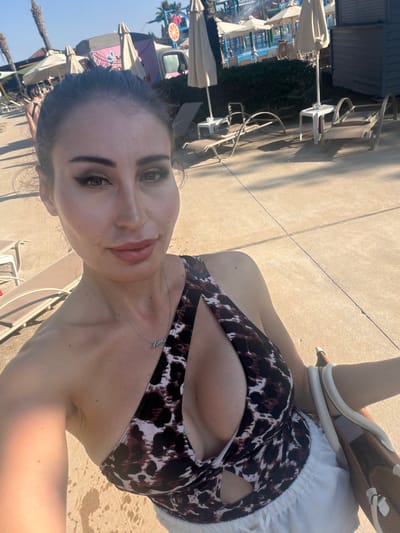 In Trachoni, Cyprus, a woman is captured in a selfie outdoors. She is positioned in the foreground, with a focus on her upper body and face. The woman is wearing a leopard-print swimsuit with a cutout detail and a silver necklace that reads "Mama". Her hair is dark and pulled back, and she has noticeable makeup, including winged eyeliner and matte lipstick. In the background, there is a swimming pool area with lounge chairs and umbrellas. A food truck with a distinctive design is also visible, along with palm trees and other vegetation. The overall setting appears to be a sunny day at a resort or water park.