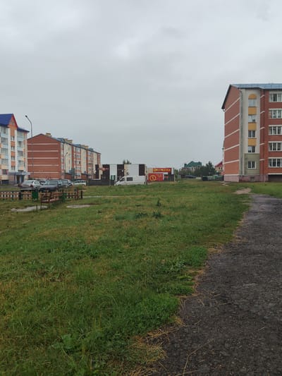 This is an outdoor, eye-level, medium-shot photograph of a residential area in Андрэеўшчынскі сельскі Савет, Belarus. The sky is overcast and cloudy. In the foreground, a dirt path is on the right side of the image, bordered by a patch of green grass and some dry grass. A small playground with a wooden fence and benches is visible in the middle ground on the left. Behind the playground are several apartment buildings. On the far left is a blue-roofed apartment building. To its right is a brick apartment building with a blue roof. Further to the right is another brick apartment building with a blue roof, and then a commercial building with a striped facade and a red awning. A white van is parked in front of the commercial building. Several cars are parked in front of the apartment buildings on the left. In the distance, more apartment buildings and some trees are visible. The overall impression is one of a relatively new or well-maintained residential area on a cloudy day.