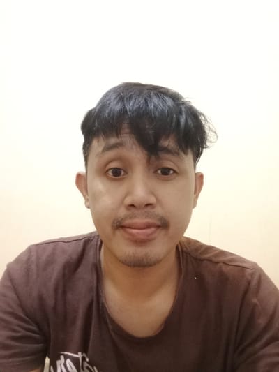 A man with short, dark hair and a slight mustache faces the camera. He has light brown skin and dark eyes. He wears a brown t-shirt. The background is a plain, off-white wall. The lighting is soft and even, illuminating the man's face. The composition is a close-up, focusing on the man's face and upper body.