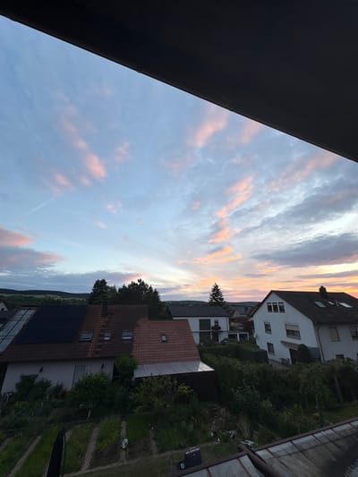A high-angle, wide shot captures a residential area in Thüngersheim, Germany, during sunset. The sky is a canvas of soft blues and pinks, with streaks of orange light radiating from the horizon. Wispy clouds catch the last rays of the sun, creating a dramatic and colorful display. In the foreground, the dark silhouette of an overhang frames the scene. Below, a cluster of houses with tiled roofs are visible. One house on the left has a large array of solar panels on its roof, while a neighboring house features a more traditional red-tiled roof. Further to the right, a white house with multiple windows stands out. Lush green trees and bushes surround the houses, and a small garden with rows of plants is visible in the lower left corner. The overall impression is one of a peaceful evening in a suburban landscape, bathed in the warm glow of twilight.