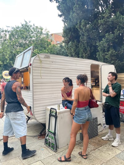 In Sant Cugat del Vallès, Spain, three individuals are gathered near a white trailer, possibly a food truck or mobile vendor. A young man in a green t-shirt and blue shorts stands near the back of the trailer, looking down and to his left. To his right, a woman in a red top and denim shorts is looking at her phone. A third person, also in a red top and denim shorts, is partially visible in the foreground, looking down. The trailer has an open service window and a sign with red lettering is visible on its side. The ground is paved with light-colored tiles. Trees are visible in the background, suggesting an outdoor setting. The overall scene appears casual and relaxed, possibly a street fair or outdoor market. The time of day appears to be daylight, and the weather seems pleasant.