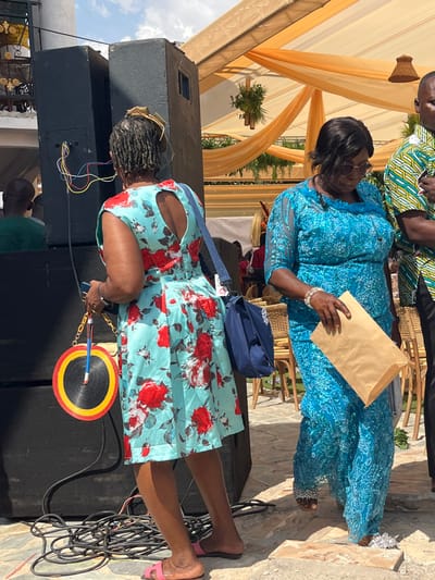 The image depicts an outdoor gathering in Kumasi, Ghana, under bright daylight. The specific location within Kumasi is not discernable.
In the foreground, a woman stands with her back mostly to the camera, wearing a light blue sleeveless dress adorned with a red floral pattern and a heart-shaped cutout on the upper back. Her hair is styled in dreadlocks, topped with a light-colored ornamental piece. She wears pink slide sandals and carries a dark blue cross-body bag over her left shoulder. In her left hand, she holds a dark-colored phone and a circular fan with black, red, and yellow concentric rings. To her immediate left are two large black speaker boxes, stacked vertically, with exposed red, blue, yellow, and green electrical wires visible near a connection point on the lower speaker. A coil of black electrical cables rests on the uneven, paved ground near her feet.
To the right, a second woman is captured in a profile view, walking or standing. She wears a bright blue, intricately patterned ensemble, possibly a dress or a top and skirt, with sheer sleeves and a ruffled neckline. Sunglasses are on her face, and a white beaded bracelet adorns her right wrist, with a silver anklet on her left ankle. She holds a light brown paper bag in her right hand. Partially visible on the far right is a man wearing a green and white patterned short-sleeved shirt.
The background features an overhead canopy made of draped orange translucent fabric, from which greenery and small woven light fixtures are suspended. Wicker chairs are visible, suggesting seating for attendees. Other blurred figures populate the background, with one man in a green shirt visible on the far left. The sky above is partly cloudy.