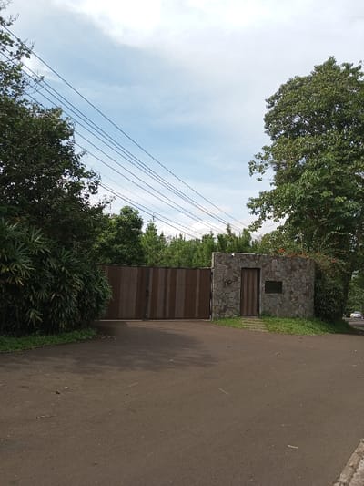 This image captures a static exterior scene in Kandenuwara, Sri Lanka, depicting the entrance to a property under a partly cloudy sky. A large, closed double gate, constructed from vertical planks with alternating light and dark brown coloration, is centrally positioned. To the right of the gate, a stone-clad gatehouse structure features a vertically-slatted wooden door, accessed by paved stepping stones, and a dark rectangular plaque affixed to the stone wall to the right of the door. Dense green foliage, including bushes and mature trees, borders both sides of the entrance. Multiple black utility wires are visible stretching diagonally across the upper left portion of the sky. The foreground consists of an asphalt driveway. Beyond the gate, a dense line of trees is visible, with a road and a white vehicle faintly discernible in the far right background. No individuals are present within the scene.