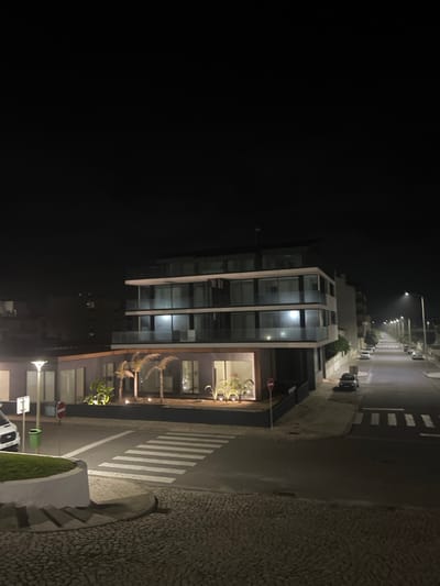 A contemporary glass-fronted building stands at night next to a deserted street illuminated by streetlights. Palm trees line the building's base, adding to its modern aesthetic. A zebra crossing lies adjacent to the building. Cars are parked alongside the street, and a textured sidewalk contrasts with the smooth asphalt. The scene conveys a sense of urban tranquility.