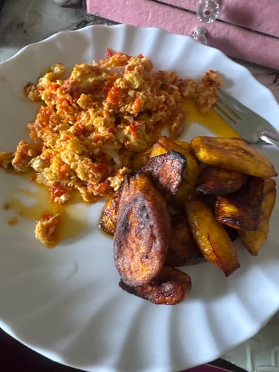 An overhead, close-up shot captures a meal served on a white, fluted plate, likely in Gbongan, Nigeria. The plate holds two main components: a generous portion of scrambled eggs mixed with finely diced red ingredients and some lighter pieces, presumably tomatoes, peppers, and onions; and a pile of deeply fried plantain slices (dodo).
The scrambled eggs exhibit a vibrant orange-yellow hue, with the red vegetable pieces integrated throughout, suggesting a flavorful preparation. A sheen of oil or sauce surrounds the base of the eggs, indicating they are freshly cooked or have been dressed. To the right of the plate, the fried plantains are golden to dark brown, some pieces showing well-caramelized edges, reflecting thorough frying.
A silver-colored metal fork is partially visible on the upper left side of the plate, with its tines resting near the egg mixture and some oil. In the blurred background, a textured, soft pink or purple fabric, possibly a tablecloth or napkin, is visible. Adjacent to it, a decorative, clear, faceted knob, likely part of furniture or decor, adds a domestic touch. The setting appears to be indoors, potentially a home kitchen or dining area, and the meal itself is typical Nigerian fare. No text is visible in the image.