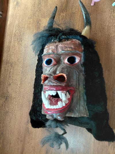 A grotesque, stylized mask is depicted hanging vertically against a light brown wooden wall with visible horizontal grain. The mask features a central face section constructed from painted, textured material, exhibiting a predominant brown hue with reddish accents around the eyes and mouth. Two large, round eyes are present, each with prominent red-rimmed eyelids, blue irises, and black pupils. A pointed, reddish-brown nose is centrally located. The mouth is wide open, revealing a row of sharp, white teeth resembling fangs, set against red gums and a dark red interior. The face is framed by thick, black, shaggy fur-like material that also extends downwards on the sides. Two horns protrude from the top of the mask; the horn on the left is dark, while the horn on the right is a lighter brown with a darker tip. A small tuft of dark gray fur hangs below the chin. The scene is located in Piperkov Chiflik, Bulgaria.