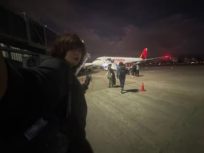 A person stands on an airport jet bridge, taking a selfie. Visible below is an Avianca airplane on the tarmac at night, with passengers disembarking. Cones are placed near the plane. The sky is dark with some cloud cover, and city lights are visible in the distance. The jet bridge structure and the person's dark clothing frame the view.