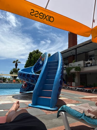 The image shows a bright, sunny day at a resort in Ayvalık, Turkey. The perspective is from a person lying down, looking up at a blue water slide shaped like a sea creature. The slide has large, dark blue circles for decoration and two red eyes peeking out from under the slide.

In the background, there is a brick building with balconies and plants. Further beyond that, there is a clear blue sky with some white clouds, and palm trees. To the left of the slide, an orange umbrella provides shade. Some lounge chairs are visible near the pool.

There are a few people in the background, some sitting on lounge chairs and others standing near the water slide. One person's feet are visible in the foreground, suggesting the photographer is lying on a lounge chair.

On the orange umbrella, the text "229X" is visible in black. The overall mood is relaxed and leisurely, typical of a vacation or a day at a pool. The lighting suggests it is daytime, likely mid-morning or afternoon given the brightness and shadows.
