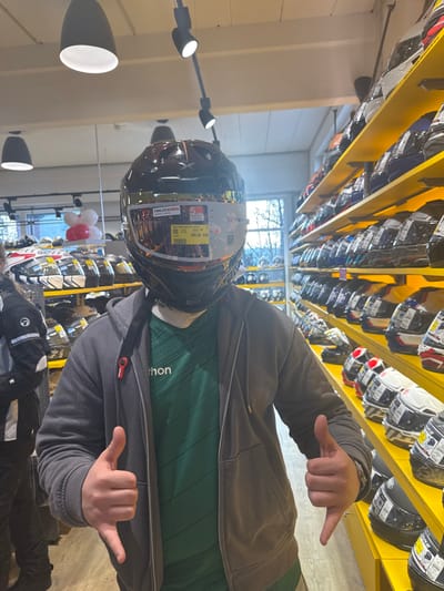 The image depicts the interior of a motorcycle gear retail store, identified as a Louis Motorrad branch in Kempten (Allgäu), Germany, characterized by distinctive yellow shelving units. A central individual, positioned facing the camera, wears a dark brown full-face motorcycle helmet with a reflective visor. A white product sticker indicating "PINLOCK" and "NEXX" branding, alongside a yellow price tag of "589.99 EUR," is visible on the helmet's visor. The individual is dressed in a dark grey zippered hoodie over a green striped t-shirt with partial text "athon" visible, and presents a "thumbs down" gesture with both hands. To the left, a second individual is partially visible, wearing a black and white motorcycle jacket and dark trousers, facing away from the camera. The store's right wall features multiple tiers of yellow shelving fully stocked with various motorcycle helmets. Additional helmets and gear are displayed on white units to the left. Overhead lighting consists of black pendant lights and multiple black track spotlights. A large window in the background provides natural light, revealing trees and an overcast sky outdoors. Two white balloons and one red balloon are attached to a display stand in the mid-ground on the left.
