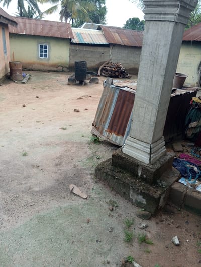 The image depicts a courtyard area in Gindin Dutse, Nigeria. In the foreground, a single sandal lies on the sandy ground. To the right, a large, unfinished concrete pillar stands, its base resting on a tiered concrete structure. Adjacent to the pillar, corrugated metal sheeting forms part of a structure, with various pieces of clothing piled nearby. Further into the scene, a pile of cut logs is visible. Behind the logs, a cylindrical metal barrel is positioned over what appears to be a makeshift fire pit constructed from stones. Another larger metal barrel stands to the left of this. The background features a building with a corrugated metal roof and a pale yellow wall, featuring a white-framed window. Several other structures with corrugated roofs are also present. The ground is predominantly sandy with patches of dry vegetation and scattered stones. The sky is overcast.