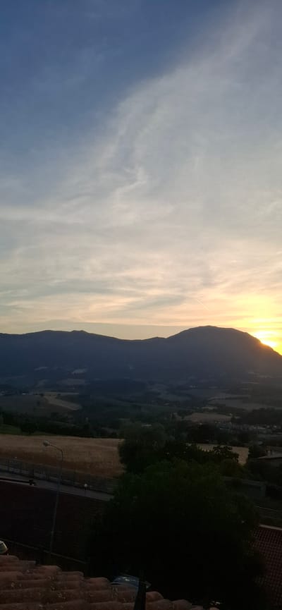 The image depicts a sunset over a rural landscape with mountains in the background. The sky is filled with wispy clouds, transitioning from a pale blue at the top to shades of orange and yellow near the horizon where the sun is setting behind a mountain range. The mountains themselves are silhouetted against the bright sky, with darker hues of blue and grey. Below the mountains, the landscape consists of rolling hills with agricultural fields, appearing to be golden in color, possibly indicating a harvest season. Scattered trees and some structures are visible on the hillsides. In the foreground, a dark mass of trees obscures the lower part of the image, and what appears to be a tiled roof is visible in the bottom left corner. A street lamp with a curved pole is also visible on a road that runs horizontally across the middle ground. The overall scene suggests a tranquil evening in a rural setting, likely in or around Camerino, Italy, given the provided context, as it captures the characteristic hilly terrain and agricultural land often found in the region. There are no people or specific actions depicted in the image.
