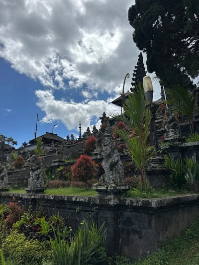 The image shows the Pura Pasar Agung Besakih temple in Rendang, Indonesia. The temple complex is composed of a series of grey stone structures covered with ornate carvings and surrounded by lush greenery, including various plants, bushes, and trees. A small palm tree is visible in the middle of the shot. The sky is partially covered by white clouds, with patches of blue visible between them.