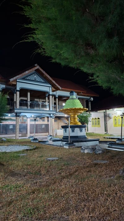 In Lhokdagang, Indonesia, at night, a building with traditional Indonesian architectural elements stands prominently. The structure features multiple levels with open balconies supported by pillars. A large, ornate golden and green fountain-like structure is positioned in the foreground, adorned with intricate carvings and topped with a green, tiered dome. Surrounding the fountain is a manicured lawn with some dried grass patches. Several lampposts are visible, casting light on the area. In the background, parts of a building with yellow-framed windows can be seen. The scene is framed by the dark branches of a large pine tree in the upper portion of the image.