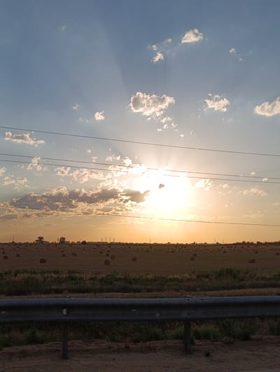 The image shows a rural landscape in the city of Z transit, Kazakhstan, at sunset. The sky is a vibrant mix of blue and orange, with rays of sunlight breaking through scattered clouds. Power lines stretch horizontally across the frame, dividing the sky from the field below. The field is dotted with numerous round hay bales, indicating an agricultural area. In the distance, a treeline is visible, along with a few structures that appear to be utility poles or pylons. A metal guardrail runs along the bottom of the image, suggesting the vantage point is from a road or elevated position. The overall scene is serene, capturing the beauty of a rural sunset.