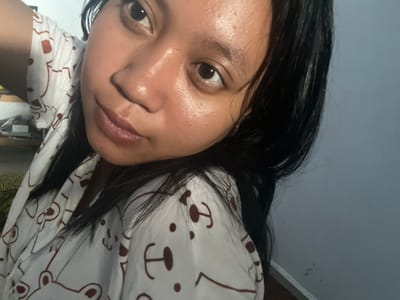 The image is a close-up, sideways selfie of a person in Jambearum, Indonesia. The person has dark hair and is wearing a white garment with a brown bear pattern. The lighting suggests it might be indoors, possibly at night or in a dimly lit room, with a gray wall in the background. The person's face is turned towards the camera, and their expression is neutral. There is no visible text in the image.