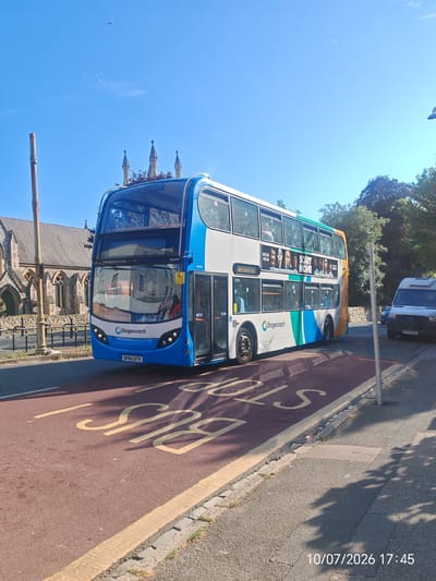 A blue and white double-decker Stagecoach bus is parked on a red bus lane, with the word "BUS" painted in yellow on the lane. The bus has a "SCARY MOVIE" advertisement on its side. In the background, a building with gothic architecture is visible, along with trees and a bright blue sky. A white van is parked on the right side of the road, and the timestamp "10/07/2026 17:45" is overlaid in the bottom right corner. The scene appears to be in Eastbourne, United Kingdom, given the presence of a Stagecoach bus, typical of UK public transport.