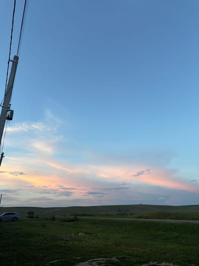 The image depicts a rural landscape under a twilight sky, not in the city of Nizhny Suyk-Su, Russia. A utility pole with wires is prominent in the left foreground, angled towards the sky. Beyond the pole, a light-colored SUV is parked on a grassy area. The midground features rolling green hills, likely farmland, with a dirt road traversing them. The sky transitions from a deep blue at the top to softer hues of pink and orange near the horizon, indicating the sun has recently set. Wispy clouds are scattered across the sky, catching the fading light. The overall scene is one of quiet, natural beauty in a rural setting.