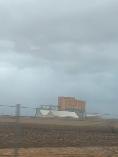 A hazy landscape features a multi-story, peach-colored building with small, evenly spaced windows adjacent to a cluster of metal silos. These industrial structures are situated near a brown, textured field that stretches into the distance. The sky is overcast, with a uniform gray color covering most of the frame. Electrical lines cut across the sky, adding a man-made element to the scene.
