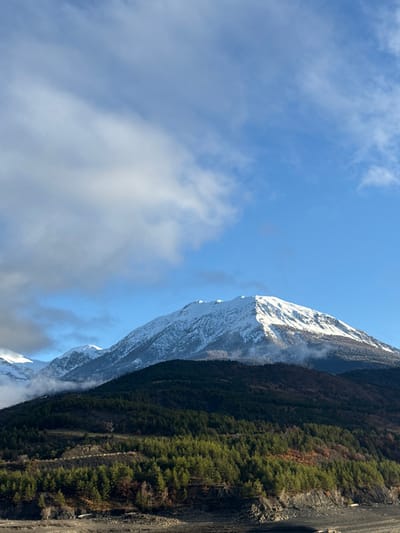 The image depicts a panoramic view of a mountainous landscape near Savines-le-Lac, France. The scene is dominated by a large, snow-capped mountain peak centrally located in the upper half, with its slopes illuminated by sunlight, creating a clear shadow line. Additional snow-covered peaks extend to the left. Below the snowline, the mountainsides are covered in dense layers of dark green coniferous forests, interspersed with areas of lighter green and reddish-brown deciduous trees, indicative of late autumn or early winter. The sky above is bright blue with scattered white and grey clouds, particularly dense on the upper left. The immediate foreground consists of dark, barren, rocky, and uneven terrain, consistent with a dry lakebed or exposed shoreline, possibly of Lac de Serre-Ponçon, with sparse scrub vegetation visible. No people, animals, or man-made structures are present.
