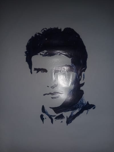 The image is a close-up of a sketch of a man's face, rendered in dark blue ink on a textured, off-white surface that appears to be some kind of fabric or tarp. The sketch is abstract and stylized, focusing on broad strokes to define the features.
The man's hair is dark and appears somewhat tousled. His left eye is clearly visible, with a strong brow and a focused gaze. The right side of his face is in shadow, with only hints of his profile suggested by the ink. The sketch includes the bridge of his nose, his lips, and his jawline, with some shading suggesting the contours of his face. He is depicted with a collared shirt or jacket, indicated by darker lines around the neck area.
The lighting in the image creates a strong highlight across the man's forehead and cheek, suggesting a light source coming from the top right. The textured surface of the background has a grid-like pattern, which is more apparent in the highlighted areas. The overall mood is serious and perhaps a little intense due to the direct gaze of the visible eye. There are no discernible signs of the time of day, weather, or specific location cues, other than the provided context of Gulbag, Uzbekistan. No text is visible in the image.