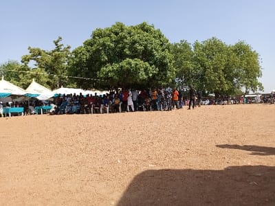 The image shows a gathering of people in Zum, Nigeria, a town located at the coordinates 10.116 latitude and 13.046 longitude. The gathering is taking place in an open, sandy field, with a large tree providing shade for a portion of the group. Several white tents are set up in the background, and the majority of people are seated on chairs or the ground. A few people are standing, interacting with each other, and some are walking away from the group. The scene suggests a social gathering, potentially a meeting or event. The environment is sunny and dry, typical of the African savanna climate.