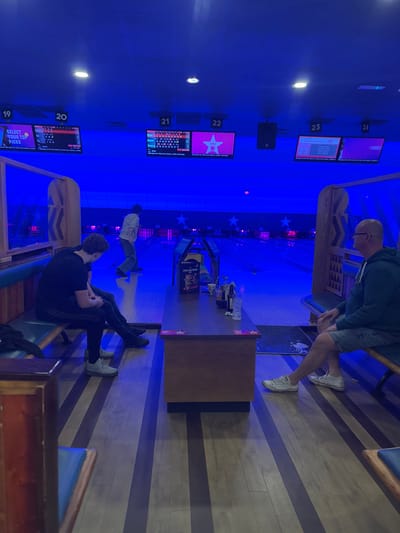 The image shows the interior of a bowling alley, located at the Hollywood Bowl, 55-59 Manchester Rd, Bradford BD1 3JF, United Kingdom. Three people are present: one person sits on a bench to the left of the frame, another person sits on a bench to the right of the frame, and a third person walks away from the camera in the distance. The person on the left wears a black hooded sweatshirt and black sweatpants, and is sitting with their legs crossed. The person on the right wears a blue and grey hooded sweatshirt and blue shorts, and is sitting with one leg crossed and the other resting on the floor. The person in the distance wears a white shirt with black detailing and black pants. There is a wooden table between the two seated people, with some items, possibly food and drink, placed on it. The room has a dark blue lighting scheme, with black and white striped flooring and several large flat-screen televisions mounted to the wall. The televisions are displaying bowling scores. The scene takes place in a bowling alley, with the bowling lanes visible in the background. The lanes are empty except for a few bowling pins. The image appears to be taken from a low angle, facing the bowling lanes.