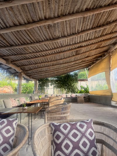 The image displays a bright, semi-outdoor seating area, likely part of a restaurant, cafe, or lounge, in Kiwengwa, Tanzania. The scene is illuminated by strong daylight, suggesting a clear day.
On the left, a prominent wall or ceiling structure is constructed from tightly woven, dried palm leaves or similar natural fibers, reinforced by rustic wooden poles. This natural material provides a textured, earthy aesthetic. Above this structure, a light-colored canvas awning with translucent sections offers partial shade or shelter.
The right side of the frame features a series of light-colored wicker chairs, some protected by clear plastic covers. Each chair is furnished with a cushion showcasing a distinctive purple and white geometric pattern. These chairs are arranged on a light-toned concrete floor, alongside simple tables, with low concrete walls defining the space.
Beyond the immediate seating area, lush green trees and foliage are visible in the background, hinting at an open-air environment or a garden setting. No people are present, lending the scene a serene and inviting atmosphere, awaiting guests. There is no visible text in the image.